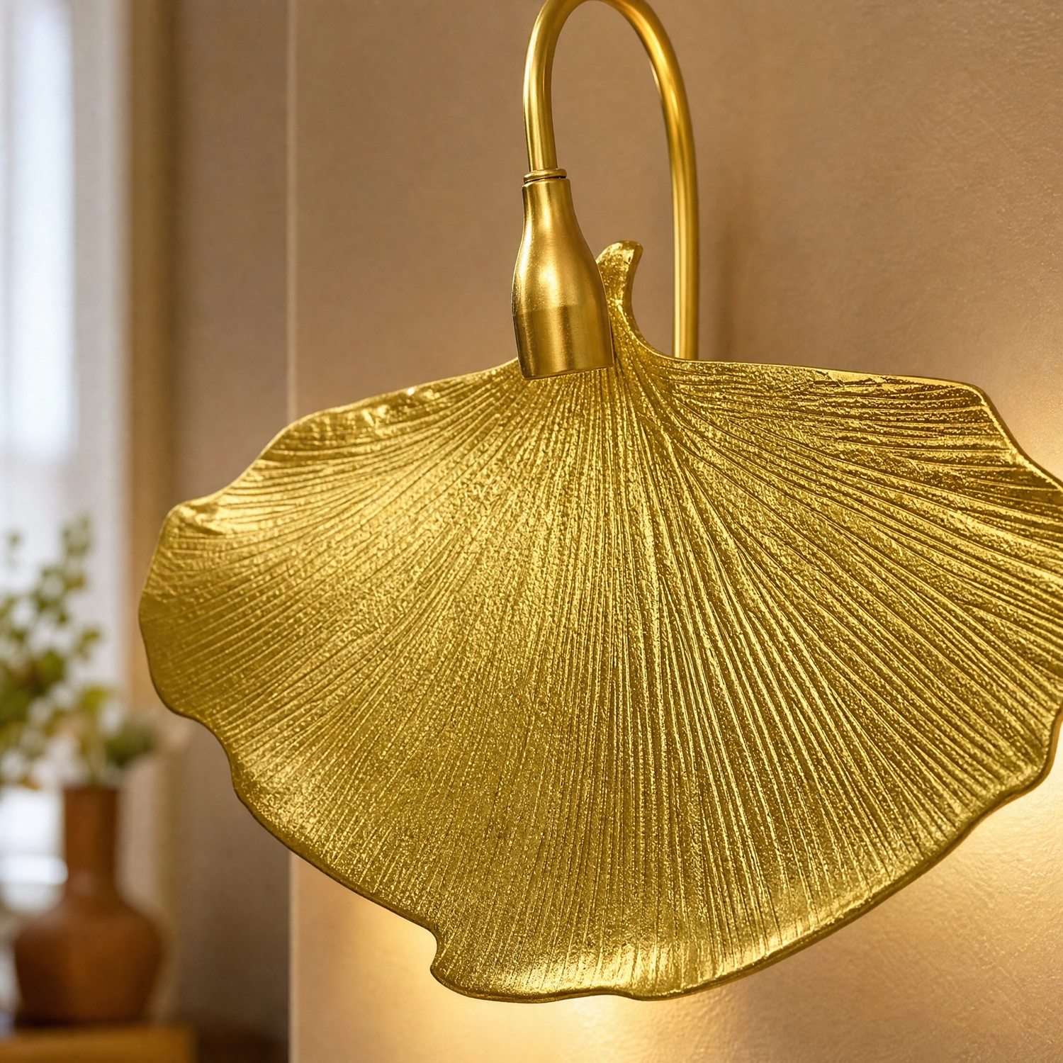 A sculptural metal wall sconce shaped like a ginkgo leaf with a textured gold finish and a curved neck.