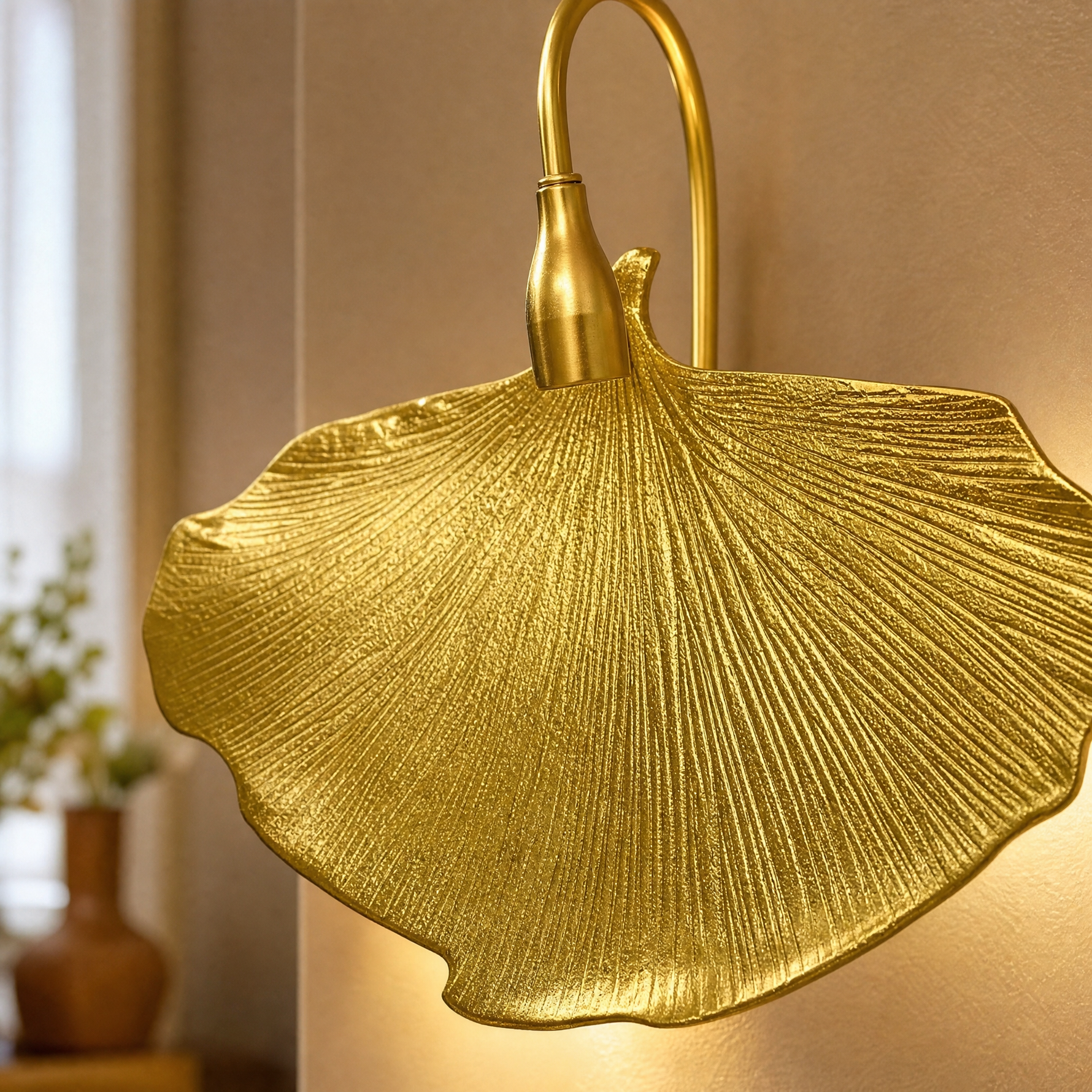 A sculptural metal wall sconce shaped like a ginkgo leaf with a textured gold finish and a curved neck.