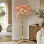A floor lamp with a blush pink ostrich feather shade and a gold, tree-trunk shaped base stands on a neutral floor.