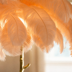 A floor lamp with a blush pink ostrich feather shade and a gold, tree-trunk shaped base stands on a neutral floor.