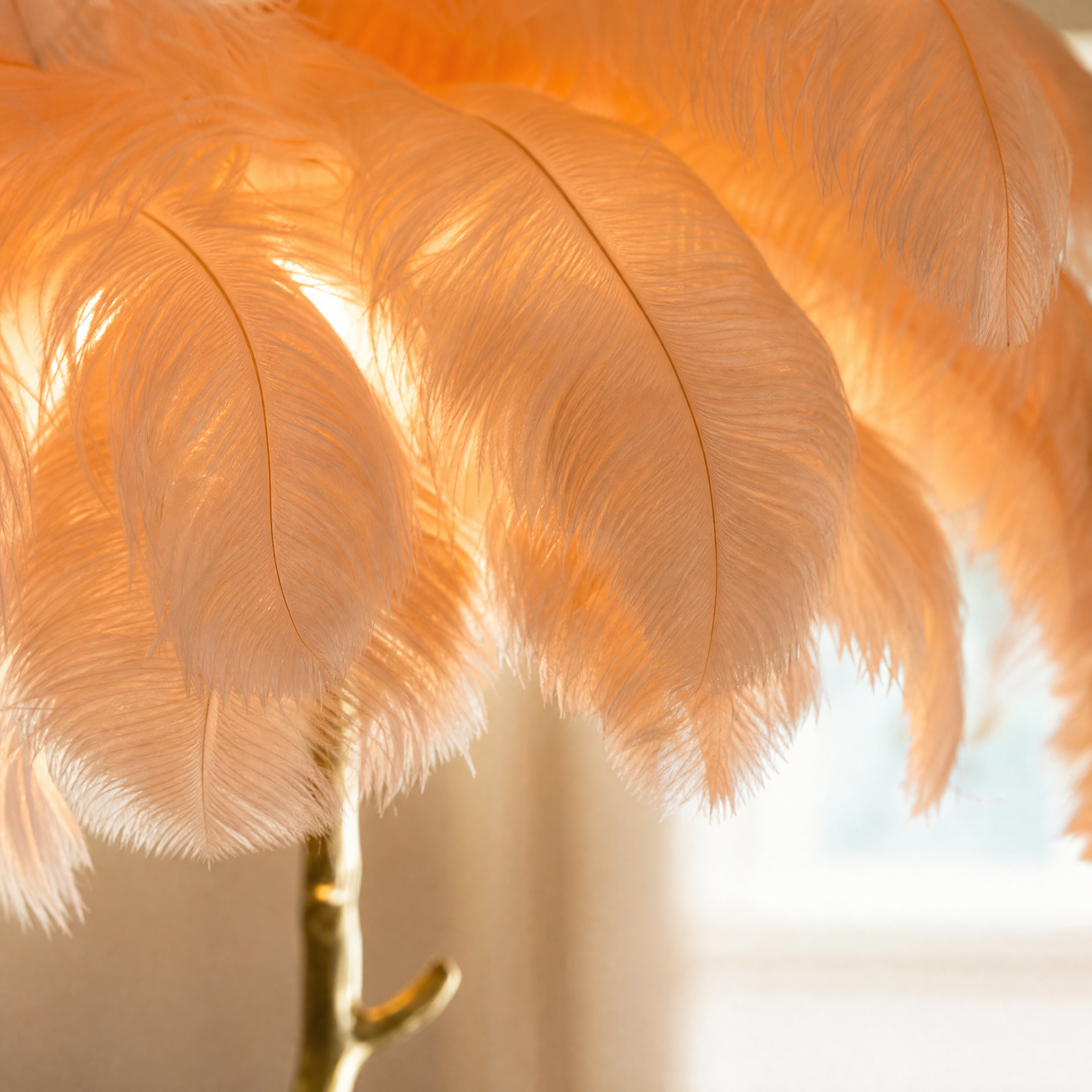 A floor lamp with a blush pink ostrich feather shade and a gold, tree-trunk shaped base stands on a neutral floor.