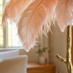 A floor lamp with a blush pink ostrich feather shade and a gold, tree-trunk shaped base stands on a neutral floor.