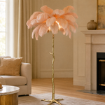 A floor lamp with a blush pink ostrich feather shade and a gold, tree-trunk shaped base stands on a neutral floor.