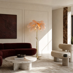 A floor lamp with a blush pink ostrich feather shade and a gold, tree-trunk shaped base stands on a neutral floor.