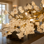 A modern organic chandelier with a warm gold branching frame and textured crystal blossom clusters hangs over a dining table.