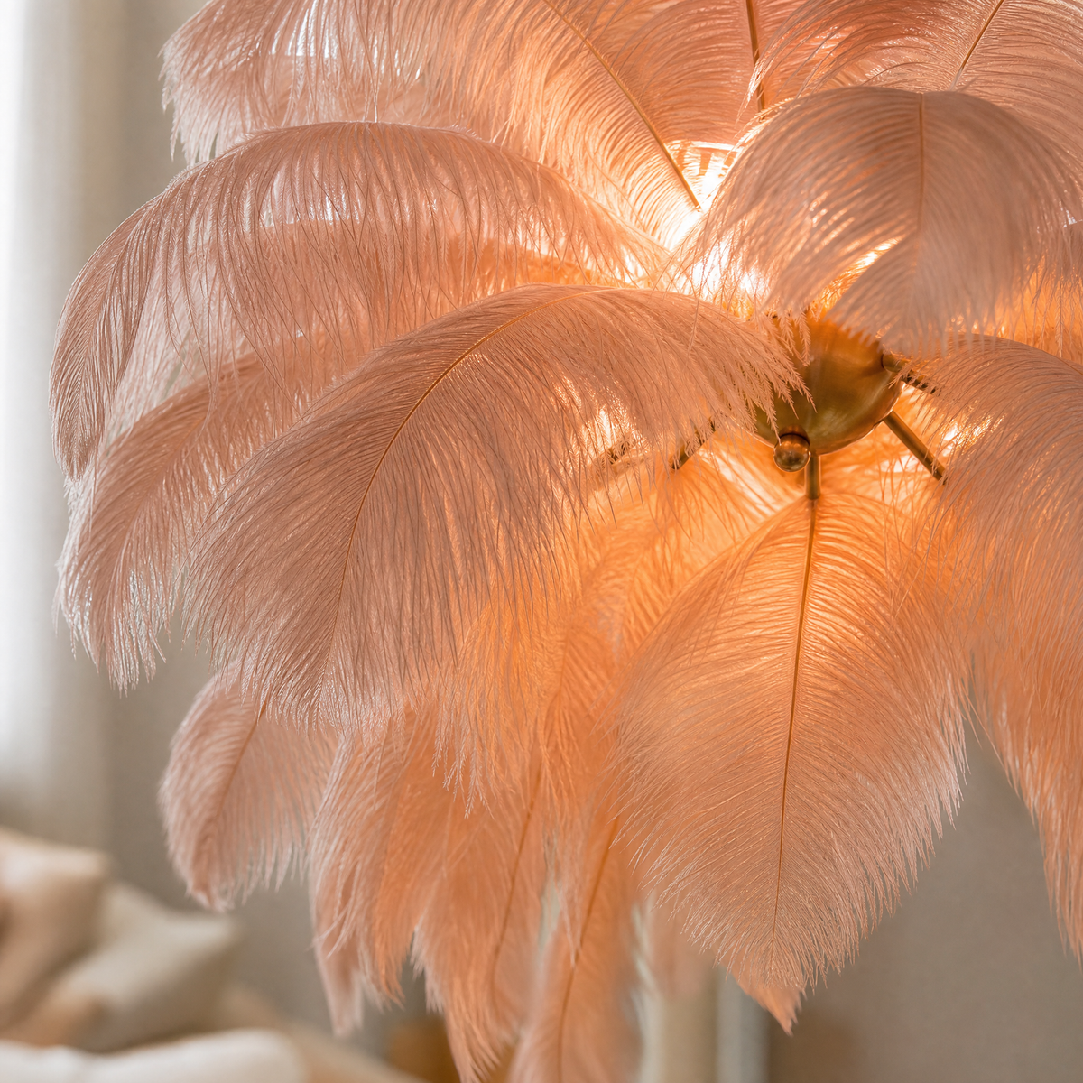 A blush pink feather pendant chandelier with a gold metal stem and circular tiered frame hangs in a bright interior space.