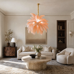 A blush pink feather pendant chandelier with a gold metal stem and circular tiered frame hangs in a bright interior space.
