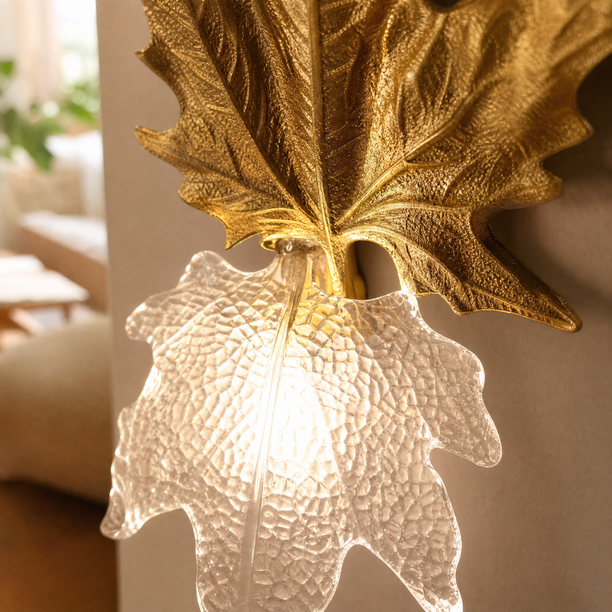 A decorative wall sconce featuring a gilded gold maple leaf backplate and a textured clear glass shade.