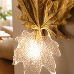 A decorative wall sconce featuring a gilded gold maple leaf backplate and a textured clear glass shade.