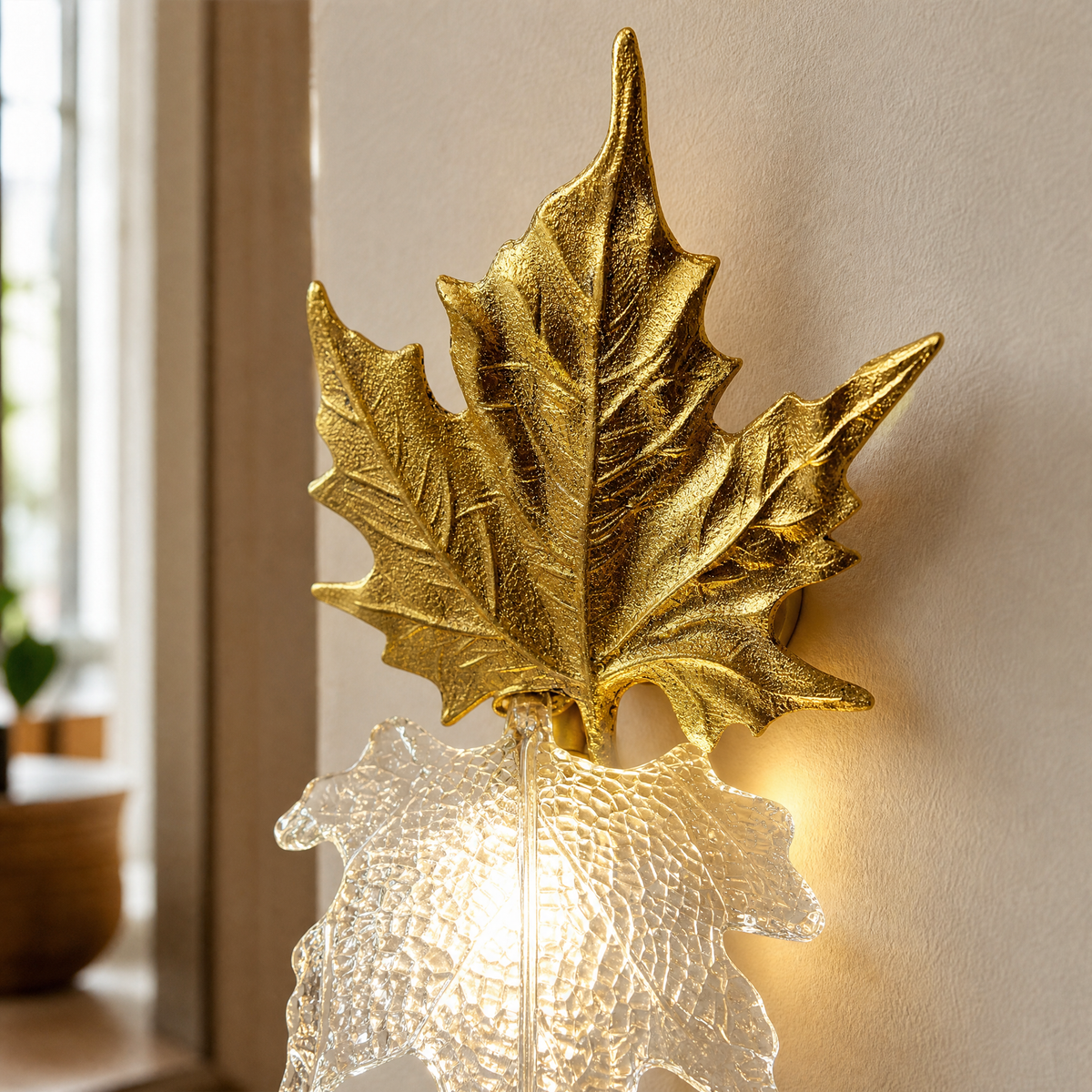 A decorative wall sconce featuring a gilded gold maple leaf backplate and a textured clear glass shade.