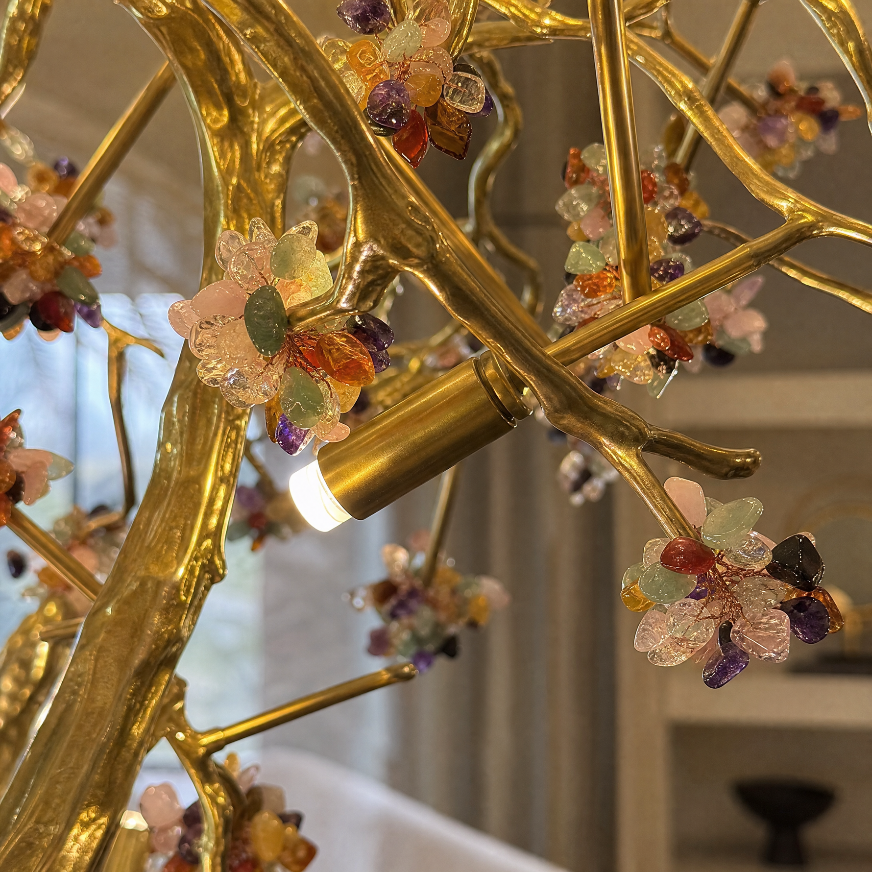 A Yiosi gold branch chandelier features a brushed brass frame with multicolored gemstone clusters and integrated downlights.