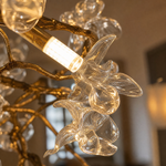 A modern organic chandelier features a warm antique brass branch frame decorated with twelve clear sculpted glass blossoms.
